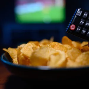 Ein Junge hält eine Fernbedienung eines Fernsehers neben einer Schüssel mit Kartoffelchips in der Hand. (Symbolbild)
