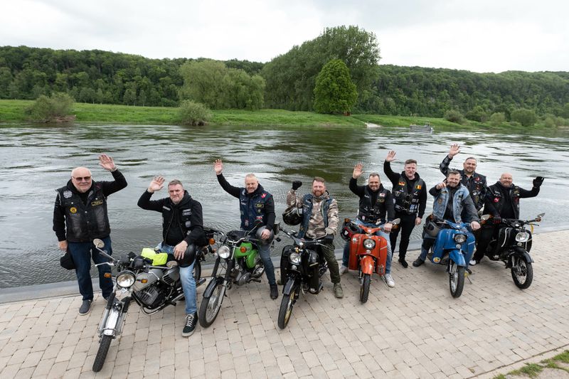 Sechs Mopedfahrer winken an einem Fluss in die Kamera.