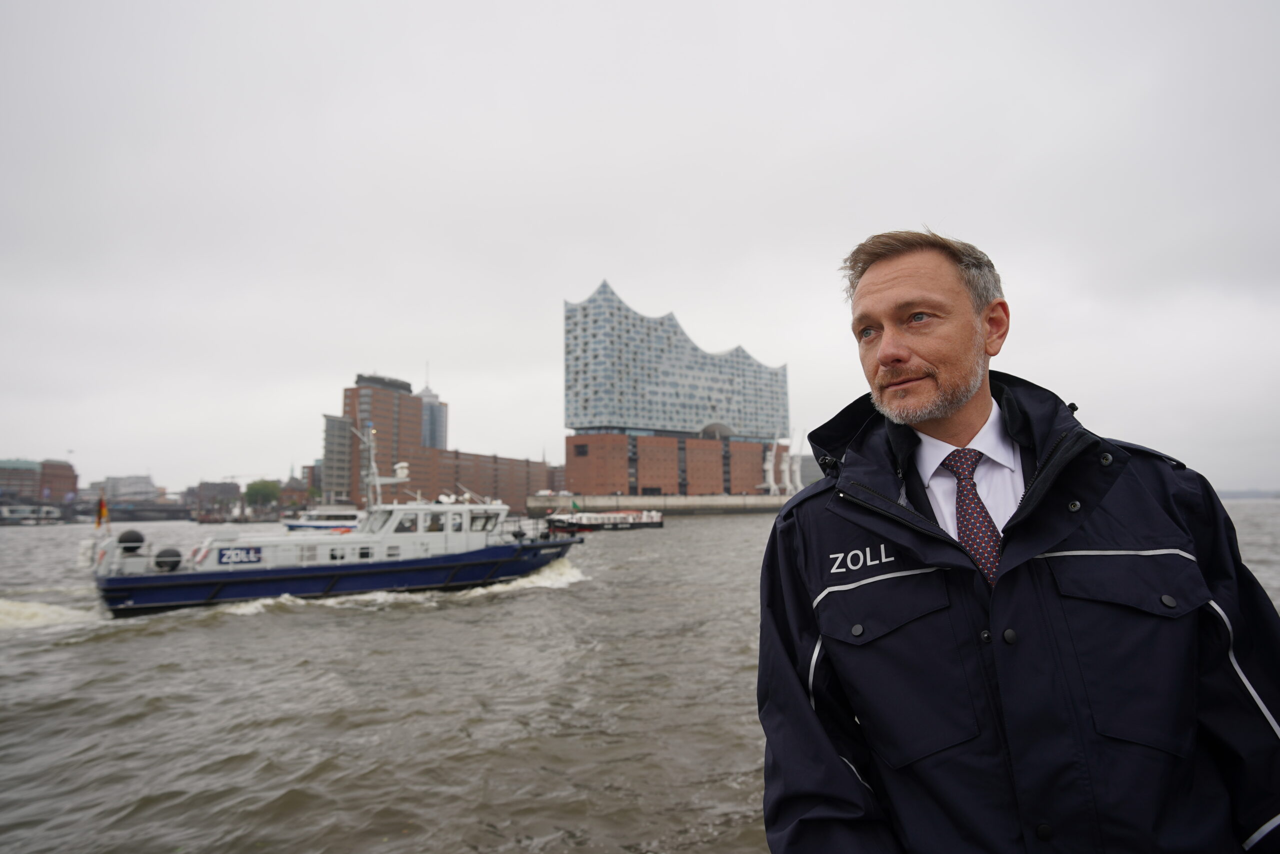 Christian Lindner (FDP), Bundesminister der Finanzen, steht an Deck des Zollbootes „Kehrwieder“ während einer Fahrt durch den Hamburger Hafen. Im Hintergrund ist die Elbphilharmonie zu sehen.