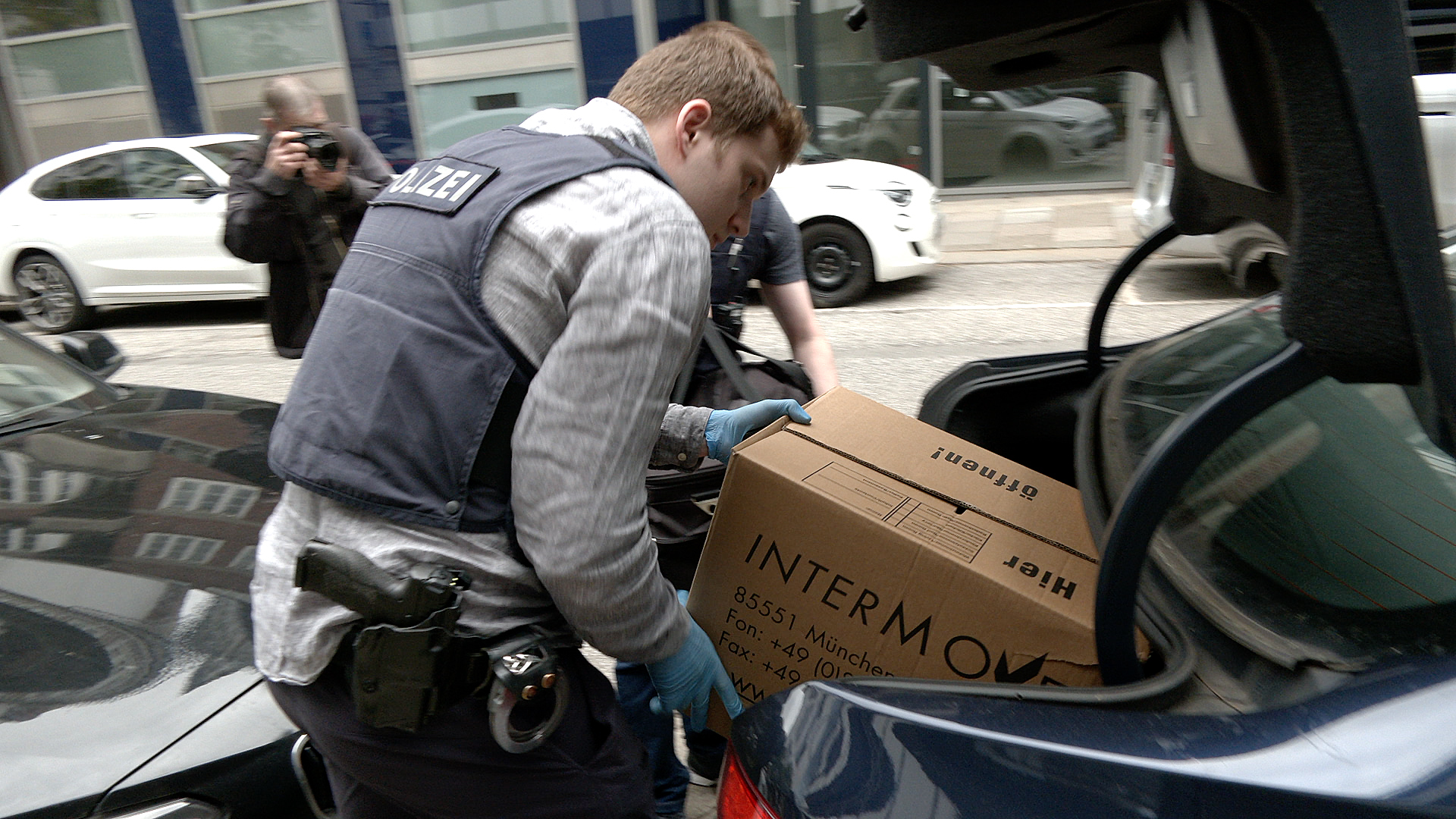 Polizisten stellen nach einer Durchsuchung einen Umzugskarton und eine Tasche in den Kofferraum eines zivilen Polizeifahrzeugs.