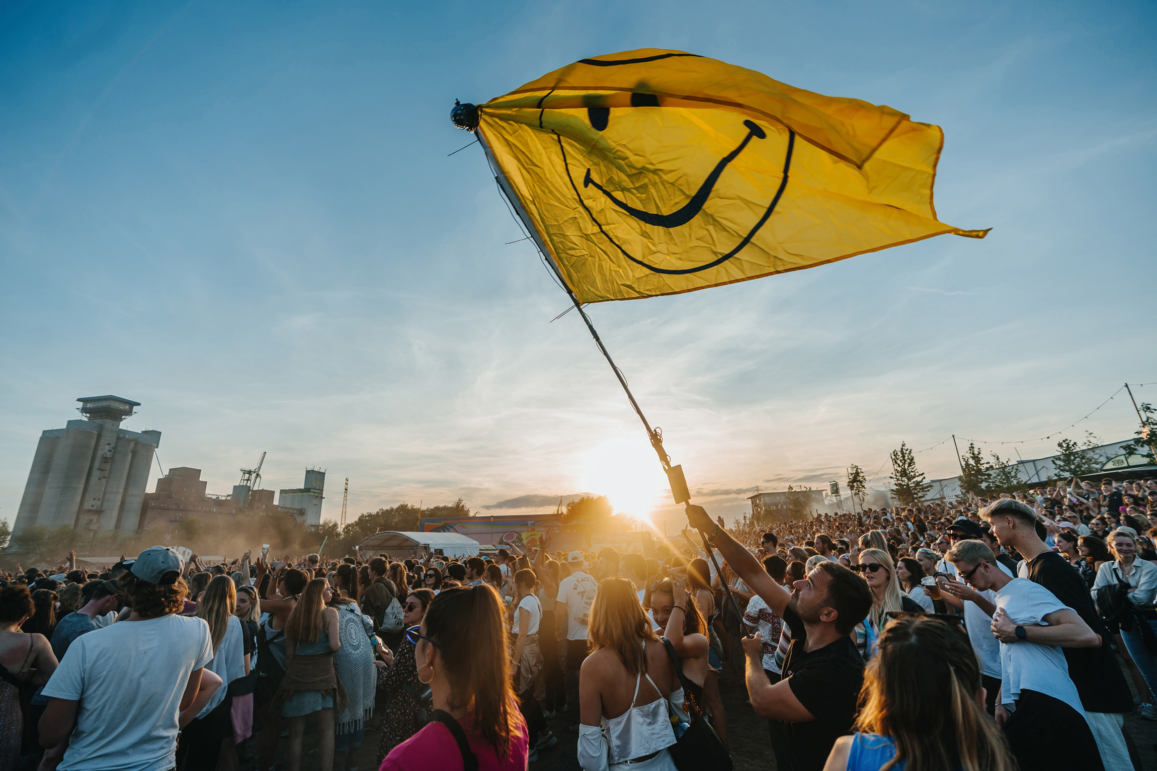 Festivalgelände, Sonnenuntergang, viele Menschen, einer Mann schwenkt vorne eine gelbe Fahne mit einem Smiley