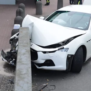 Ein weißer Maserati Ghibli steht mit zerdrückter Front an einer Mauer, im Hintergrund Feuerwehrleute und Polizisten