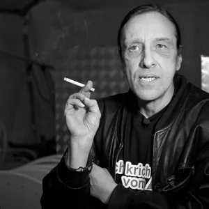 Ein Schwarzweiß-Foto von Arno Dübel. Auf seinem Shirt steht „Ich krich vom Amt“, er hält eine Zigarette in der Hand