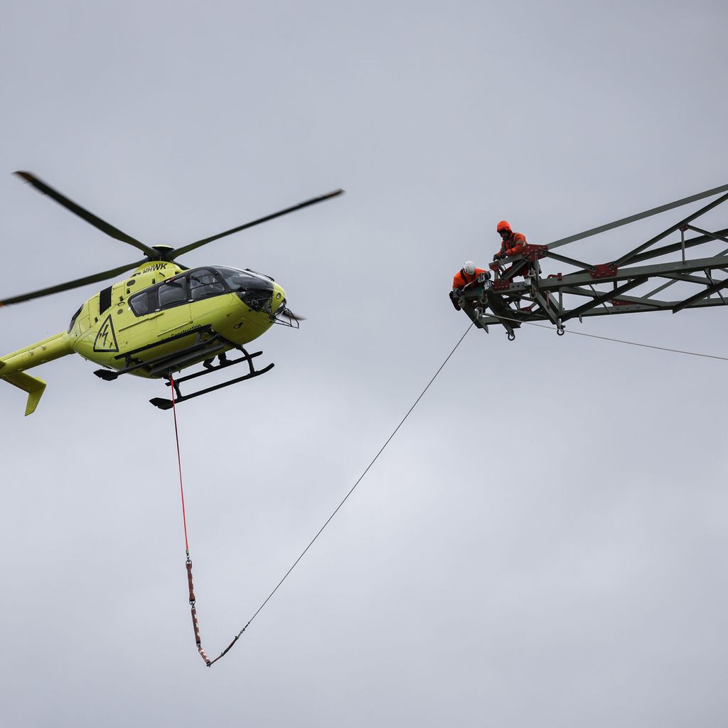 Hubschrauber fliegt auf Höhe eines Strommastes, auf dem ein Arbeiter steht. Ein Seil verbindet beide Objekte