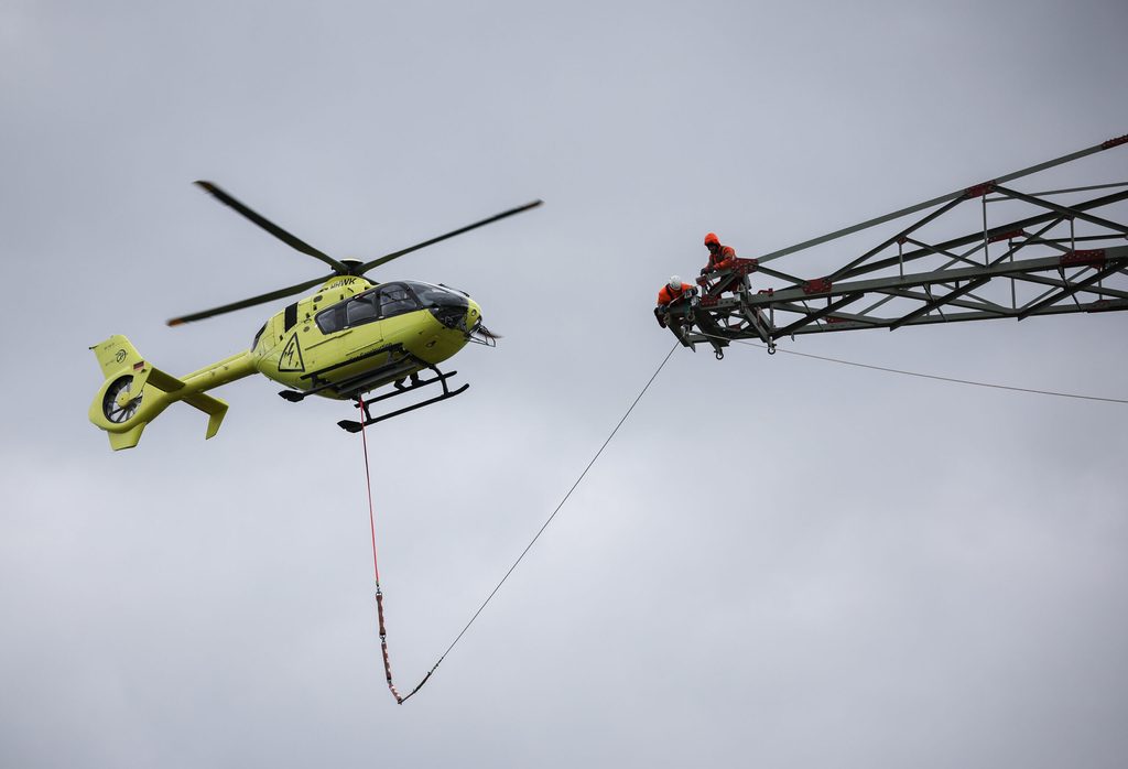 Hubschrauber fliegt auf Höhe eines Strommastes, auf dem ein Arbeiter steht. Ein Seil verbindet beide Objekte