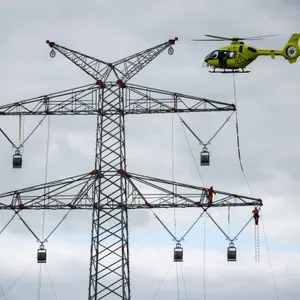 Hubschrauber fliegt über Strommast auf dem zwei Menschen stehen, am Hubschrauber ist ein Seil befestigt.
