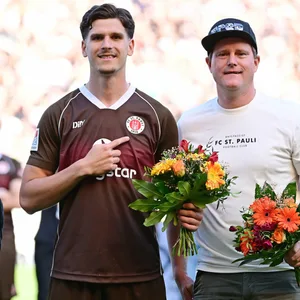 Igor Matanovic weist auf das St. Pauli-Logo, daneben Oke Göttlich, beide mit Blumenstrauß.