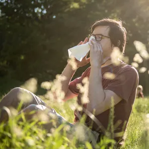 Ein Mann sitzt bei Sonnenschein auf einer Wiese und putzt sich die Nase mit einem Taschentuch.