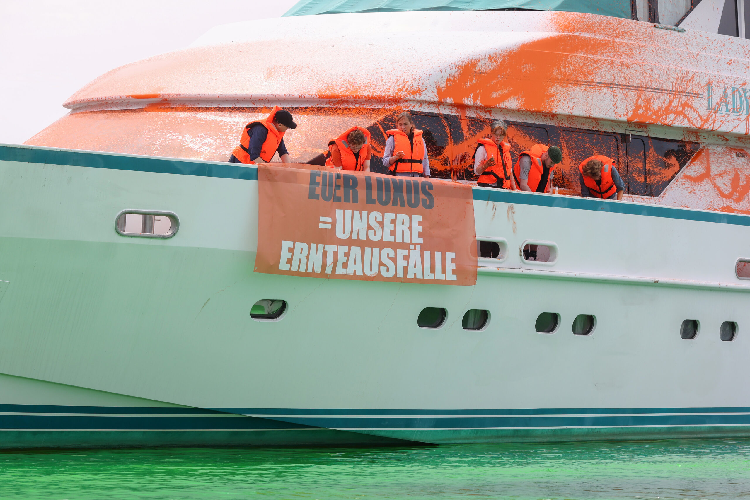 Teilnehmer:innen einer Protestaktion stehen mit Transparenten auf einer Yacht im Hafen von Neustadt. Auf einem Transparent ist die Aufschrift „Euer Luxus = Unsere Ernteausfälle“ zu sehen.