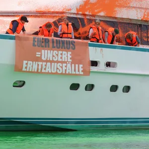 Teilnehmer:innen einer Protestaktion stehen mit Transparenten auf einer Yacht im Hafen von Neustadt. Auf einem Transparent ist die Aufschrift „Euer Luxus = Unsere Ernteausfälle“ zu sehen.