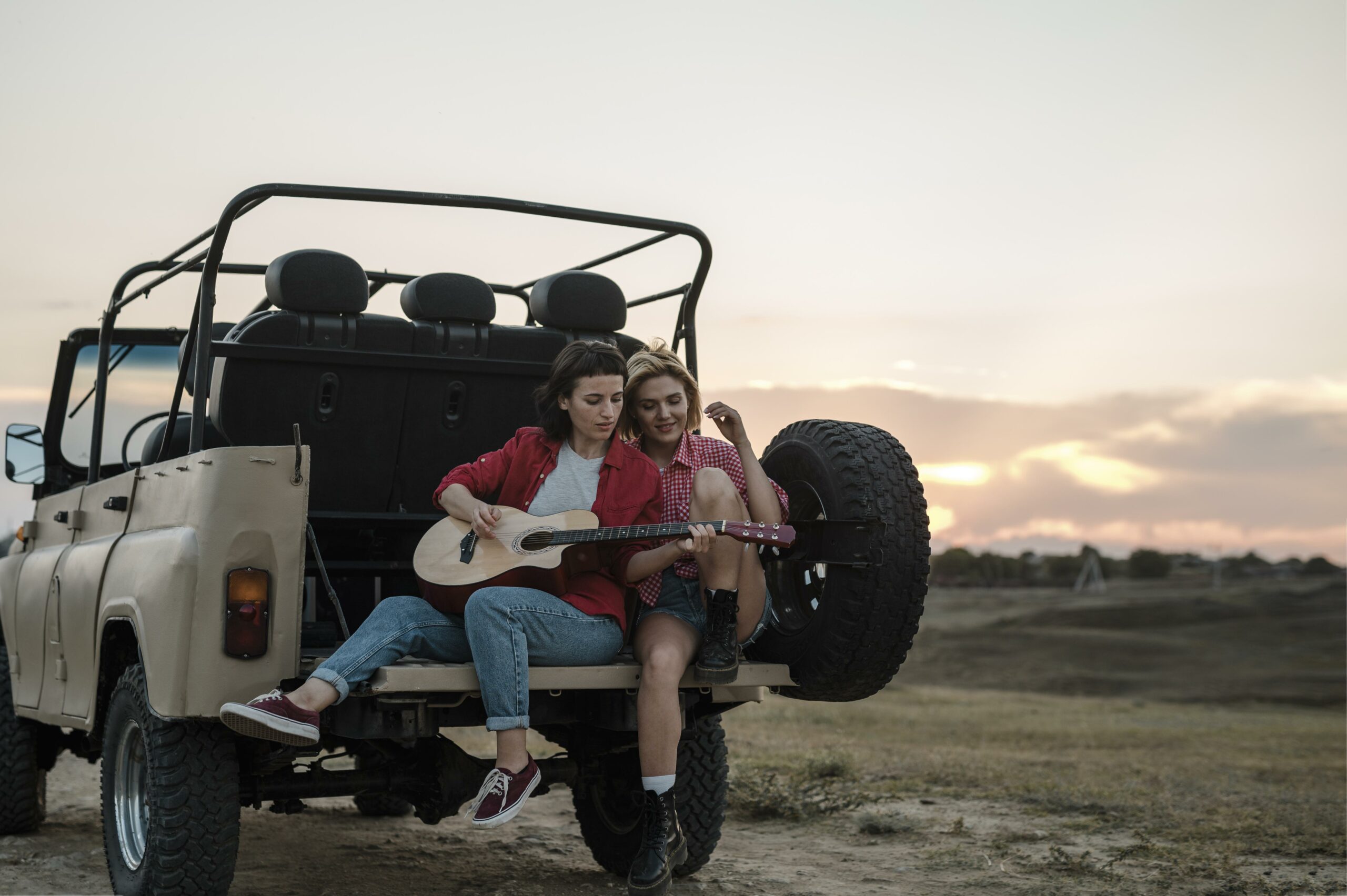 Zwei Frauen sitzen auf einem Fahrzeug, das im Sonnenuntergang auf einem Feld steht, eine Frau spielt dabei Gitarre. (Symbolbild)