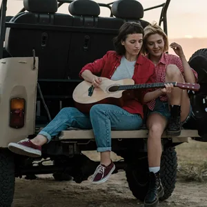 Zwei Frauen sitzen auf einem Fahrzeug, das im Sonnenuntergang auf einem Feld steht, eine Frau spielt dabei Gitarre. (Symbolbild)