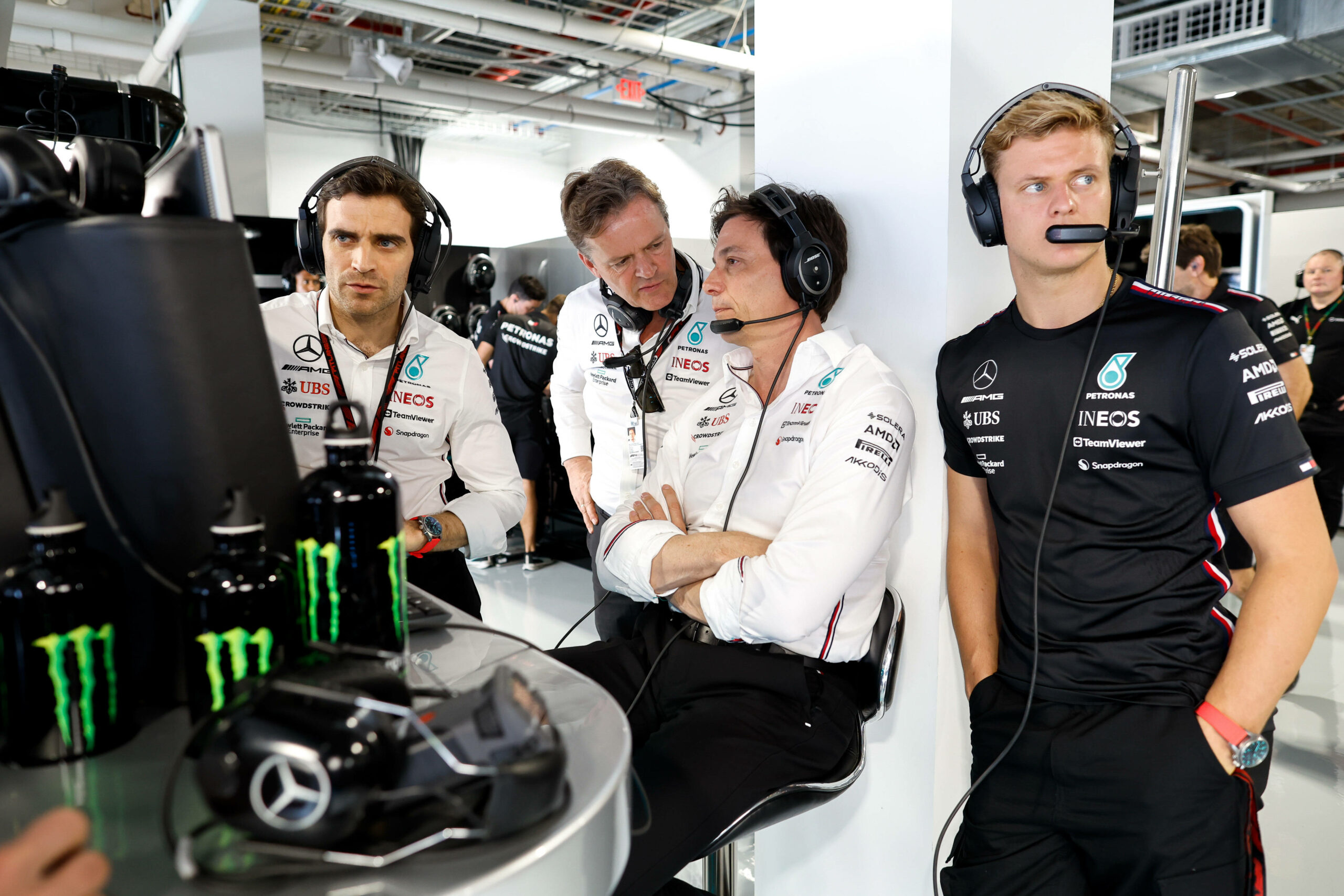 Mick Schumacher in der Garage von Mercedes mit einem Headset auf. Im Hintergrund unterhält sich Teamchef Toto Wolff mit Markus Schäfer.