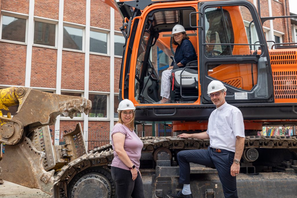 Die drei Personen umringen einen Bagger vor dem alten Schulgebäude.