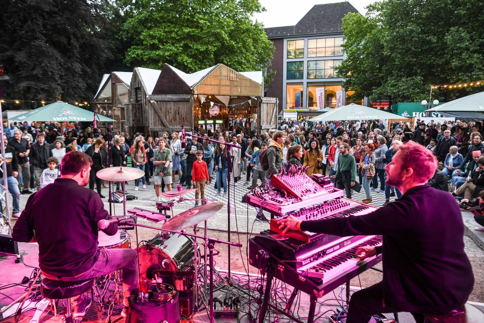 Im Vordergrund eine Bühne mit Musikern, hinten stehendes Publikum