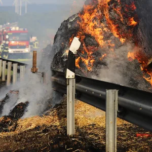 Aufnahme des brennenden Anhängers, mit Feuerwehrleuten im Hintergrund.