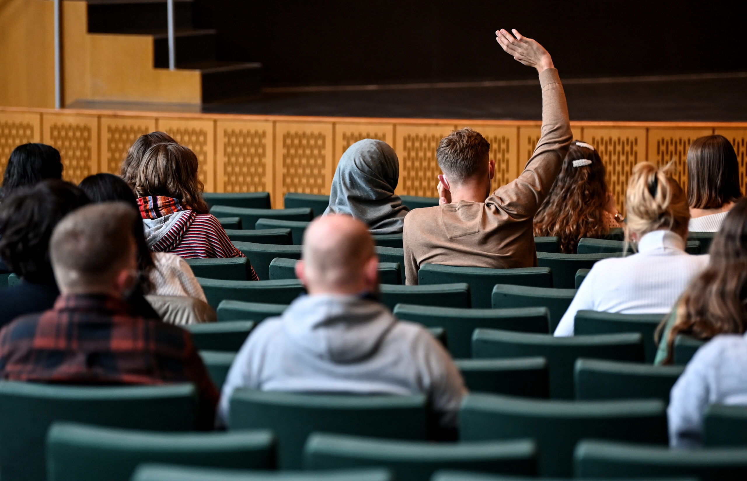 Studentinnen und Studenten in einem Hörsaal. Sie sind von hinten zu sehen und ein Student hebt die Hand.