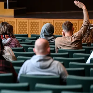 Studentinnen und Studenten in einem Hörsaal. Sie sind von hinten zu sehen und ein Student hebt die Hand.
