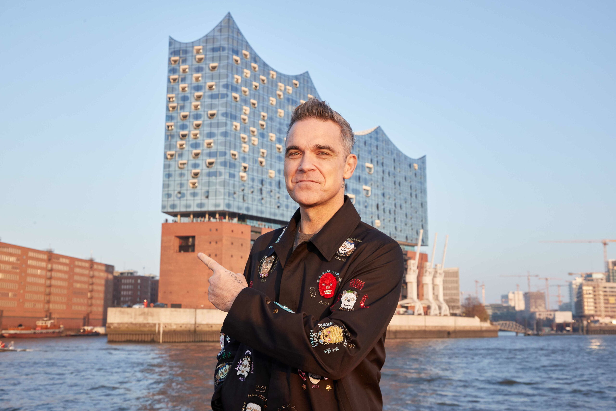 Robbie Williams, britischer Sänger und Songwriter, steht während eines Fototermins auf einer Barkasse im Hamburger Hafen. Im Hintergrund ist die Elbphilharmonie zu sehen. (Archivbild)