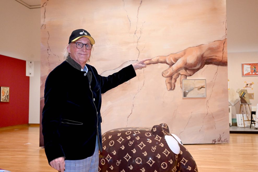Der Komiker und Künstler Otto Waalkes steht nach dem Presserundgang "Otto die Ausstellung" von Otto Waalkes im Buchheim Museum neben einer Figur eines Ottifanten und zeigt dabei auf die gemalte Hand Gottes.