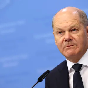 Olaf Scholz schaut bedröppelt drein, vor ihm ein Mikrofon, hinter ihm eine blaue Pressewand.