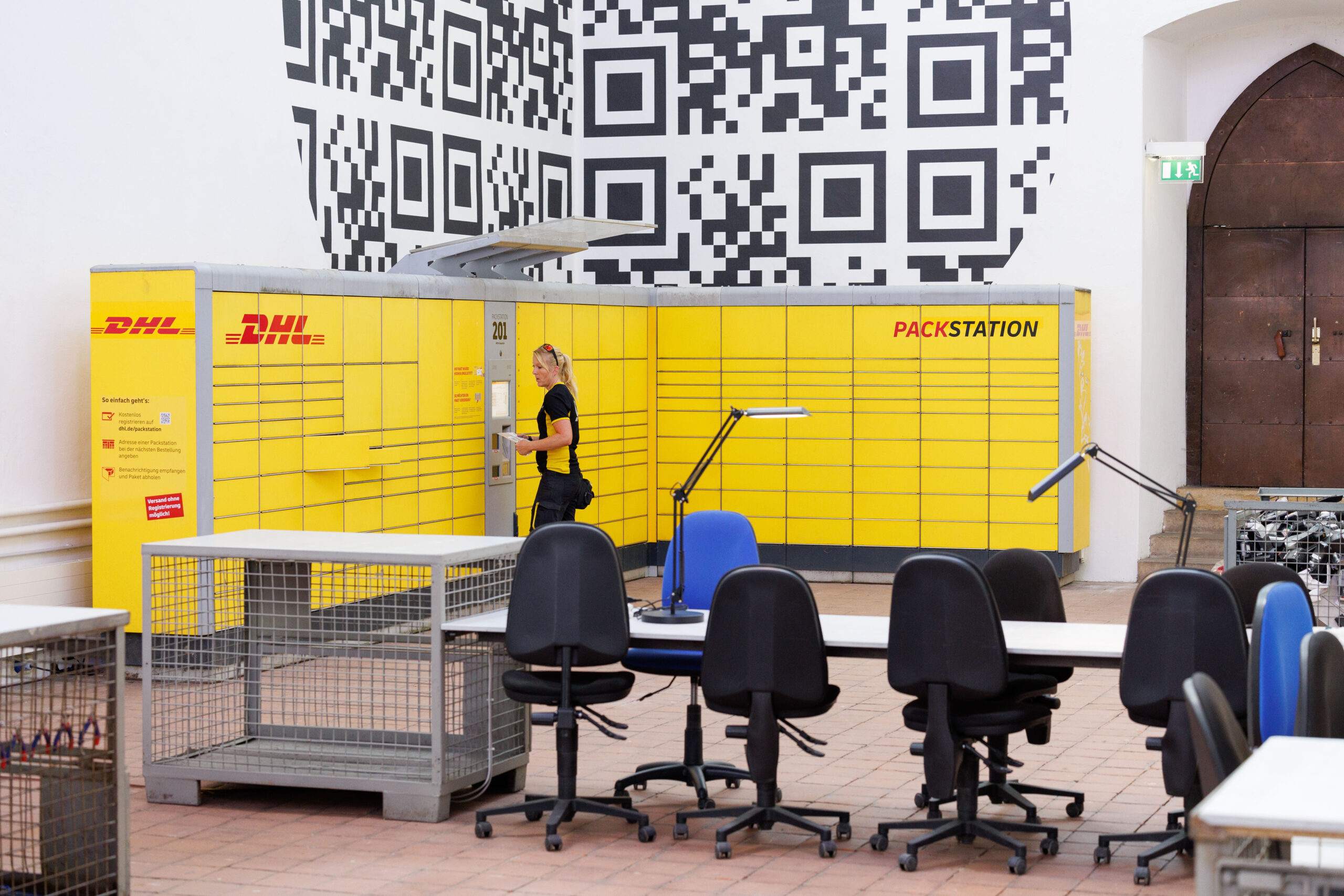Eine DHL-Pakstation steht vor einer Wand mit einem großen QR-Code.