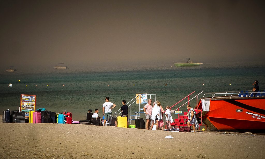 Vor einem dunklen Himmel stehen Touristen mit ihrem Gepäck am Strand.