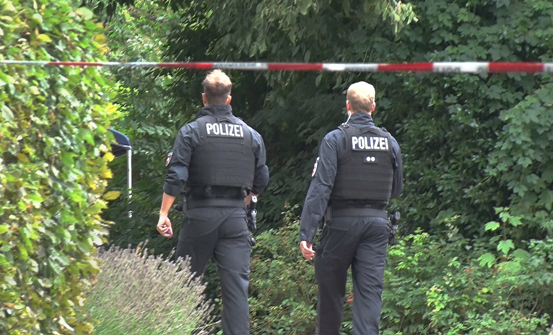 Zwei Polizisten gehen hinter einer Absperrung zu dem Gebüsch, in dem die Spaziergängerin die Leiche fand.