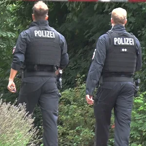 Zwei Polizisten gehen hinter einer Absperrung zu dem Gebüsch, in dem die Spaziergängerin die Leiche fand.