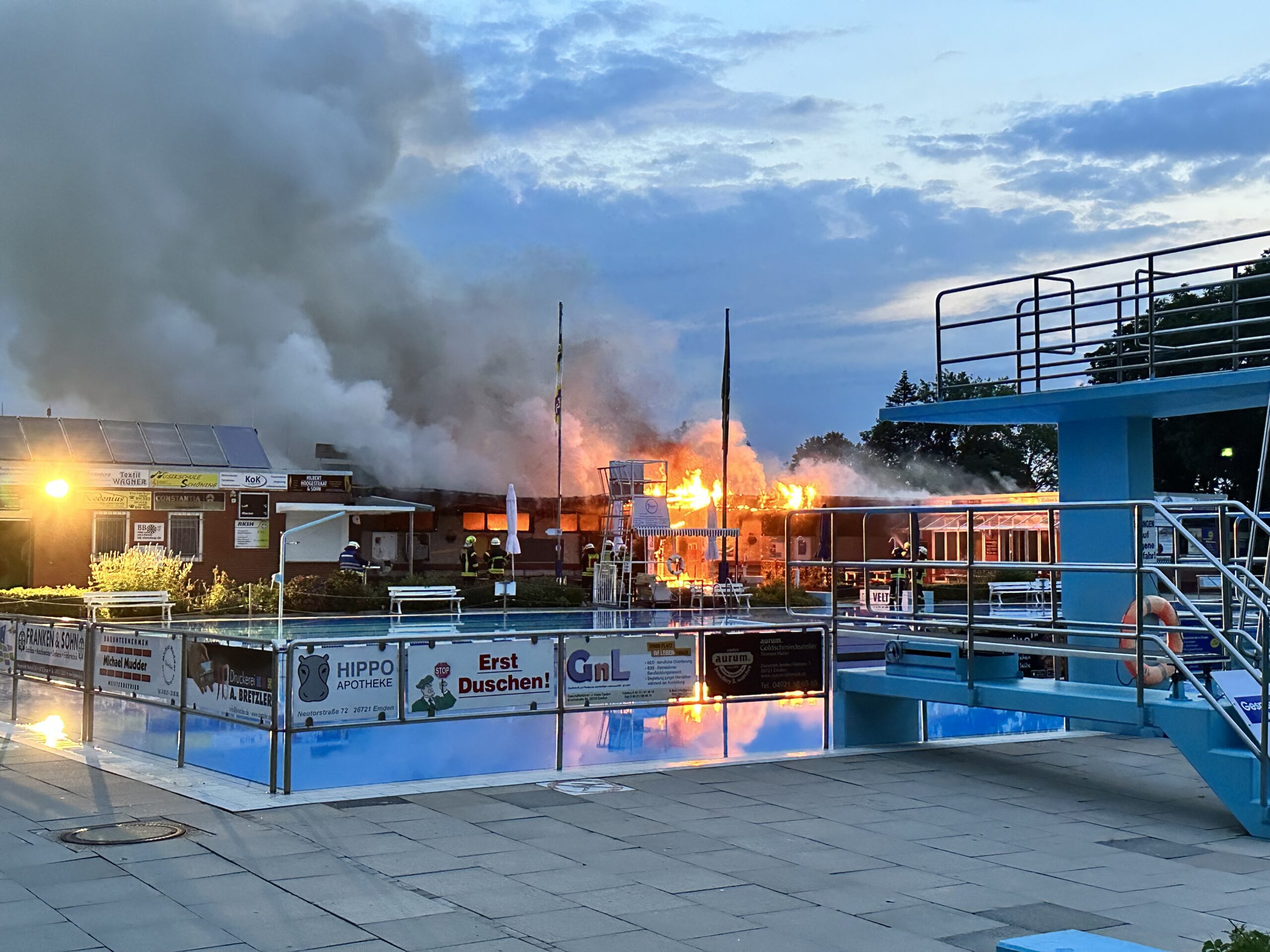 Freibad-Backen mit Sprungturm, Gebäude dahinter steht in Flammen.