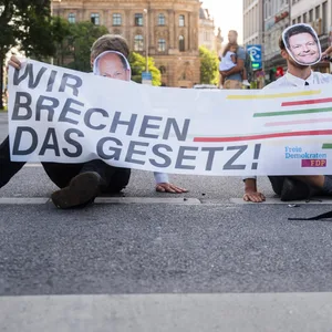 Zwei Personen der Letzten Generationen blockieren eine Straße. Jeweils eine Hand ist an die Fahrbahn geklebt, mit der anderen halten sie ein Banner „Wir brechen das Gesetz!“, mit den Logos der Regierungsparteien. Ein Teilnehmer trägt eine Olaf-Scholz-Maske, der andere eine Robert-Habeck-Maske.