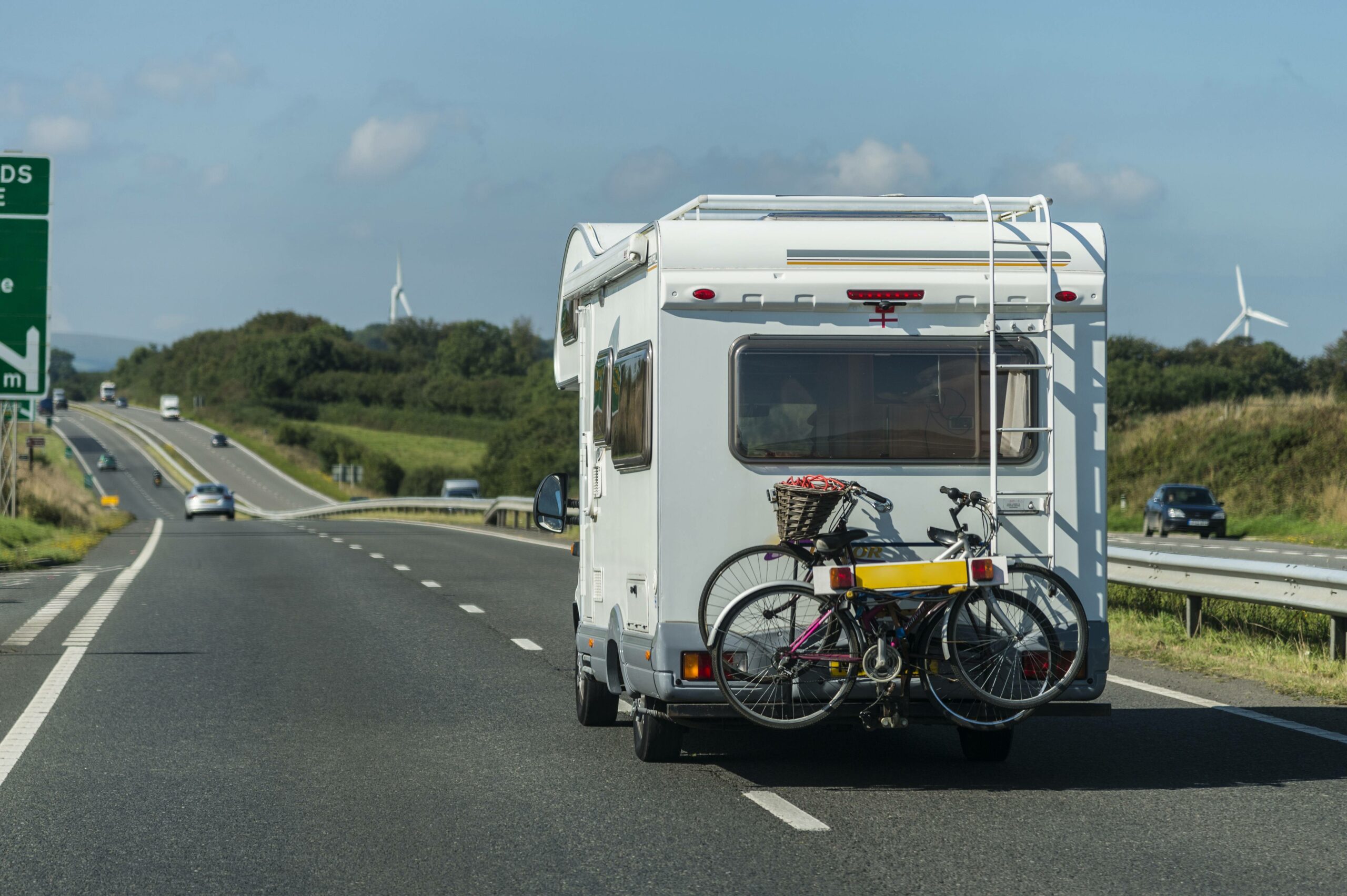 Ein Wohnmobil auf einer Schnellstraße von hinten. Im Hintergrund grüne Wiesen, Bäume und Windräder.