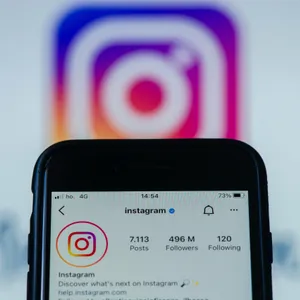 Im Hintergrund ist ein unscharfes Instagram-Logo, davor hält jemand ein Smartphone mit der geöffneten Instagram-App in die Kamera.