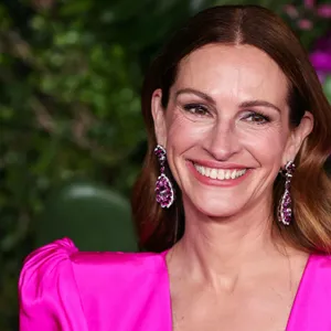 Schauspielerin Julia Roberts in einem pinken Kleid mit großen Uhrringen, lachend, hinter ihr ist eine Wand aus Pflanzen.