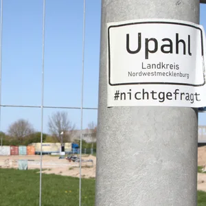 Hinter einem Bauzaun sind mehrere bunte Container und eine Sandfläche sichtbar, im Vordergrund rechts ein Laternenpfahl mit Aufkleber „Upahl – Landkreis Nordwestmecklenburg – #nichtgefragt“