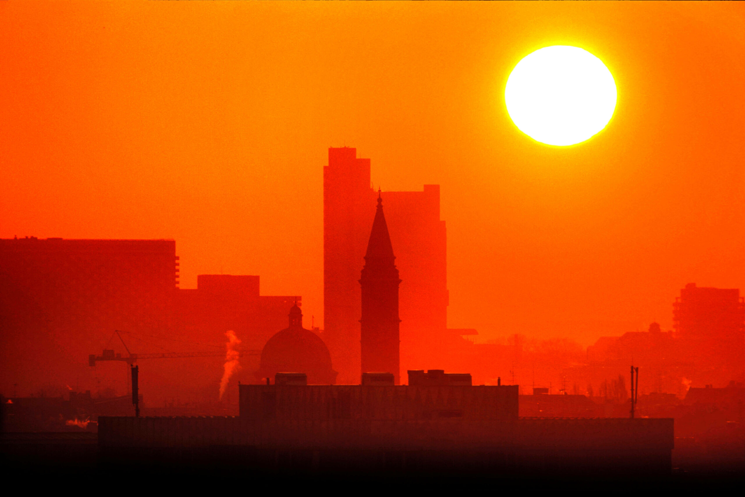 Die Skyline einer Stadt, darüber steht die gleißend helle Sonne als großer Ball.