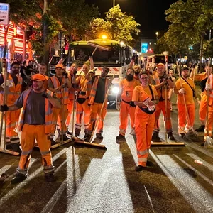 Mitarbeitende in Arbeitskleidung posieren mit Besen auf der dunklen Straße.