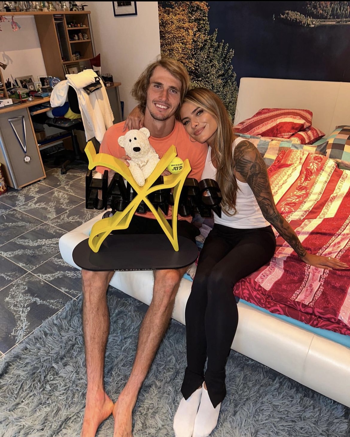 Alexander Zverev mit Pokal und Sophia Thomalla sitzen auf dem Kinderbett von Zverev und lächeln in die Kamera.