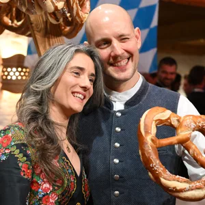 Oliver Petszokat mit Frau Pauline bei einer Weißwurst-Party in Österreich: Oli P. hat eine große Brezel in der Hand, hinter ihnen ist eine blau-weiße Flagge.