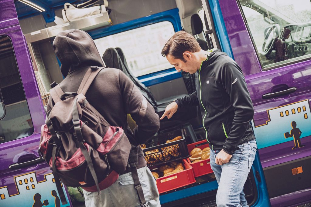 Ein Helfer steht am Mitternachtsbus und gibt einem Obdachlosen ein Brötchen