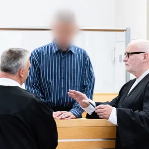 Im Prozess zum versuchten Mord mit Rattengift: Angeklagter in der Mitte, rechts und links zwei Männer in schwarzen Gerichts-Kutten