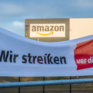 Verdi-Banner im Vordergrund mit Aufschrift „Wir streiken“, Amazon Logistikcenter mit Aufschrift „amazon“ im Hintergrund.