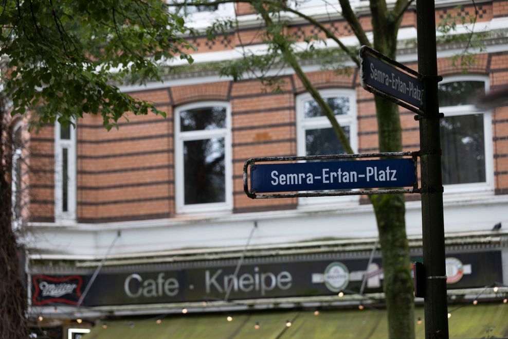 Man sieht ein blaues Straßenschild mit der Aufschrift „Semra-Ertan-Platz“, dahinter ein Wohnhaus, im Erdgeschoss das Café Miller.