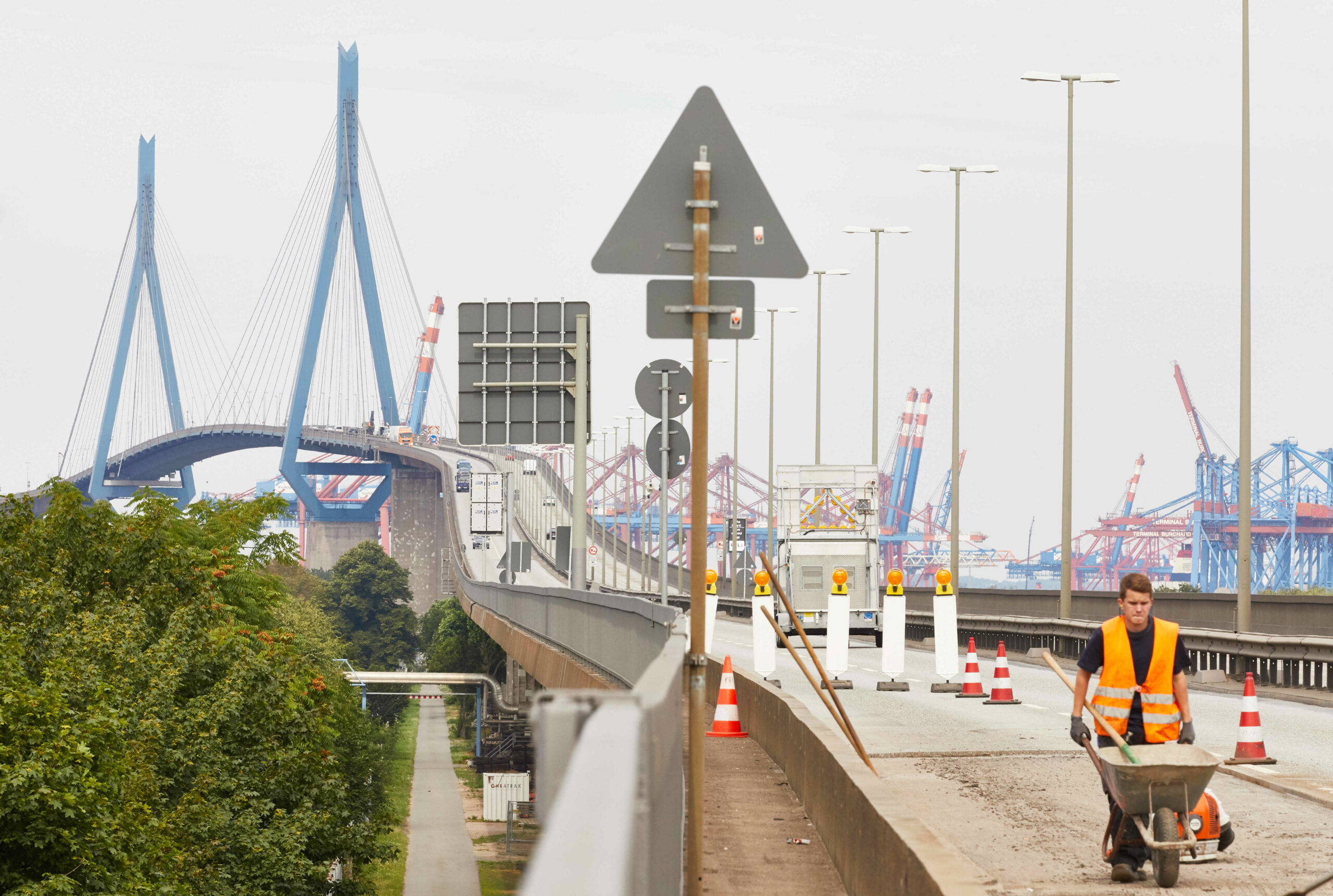 Man sieht eine große Brücke, die Fahrbahn ist mit Hütchen abgesperrt und ein Bauarbeiter läuft mit einer Schubkarre entlang.