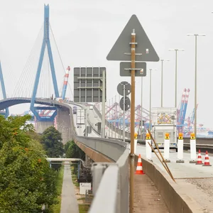 Man sieht eine große Brücke, die Fahrbahn ist mit Hütchen abgesperrt und ein Bauarbeiter läuft mit einer Schubkarre entlang.