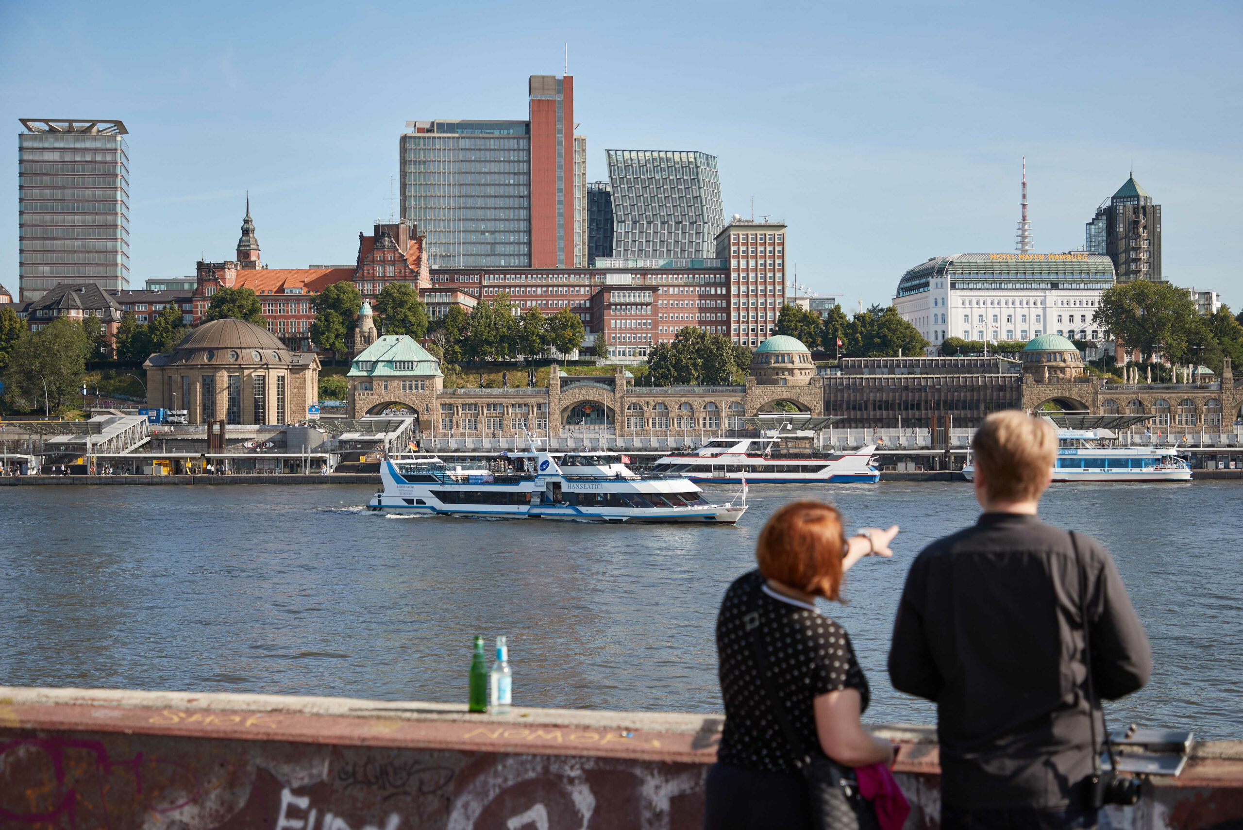 Zwei Menschen betrachten bei Sonnenschein im Hamburger Hafen die Stadt. Im Hintergrund sind der Alte Elbtunnel und die Landungsbrücken zu sehen.