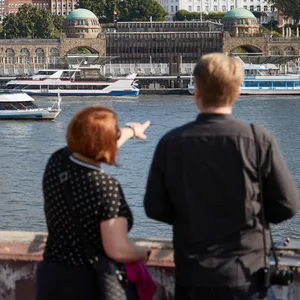 Zwei Menschen betrachten bei Sonnenschein im Hamburger Hafen die Stadt. Im Hintergrund sind der Alte Elbtunnel und die Landungsbrücken zu sehen.