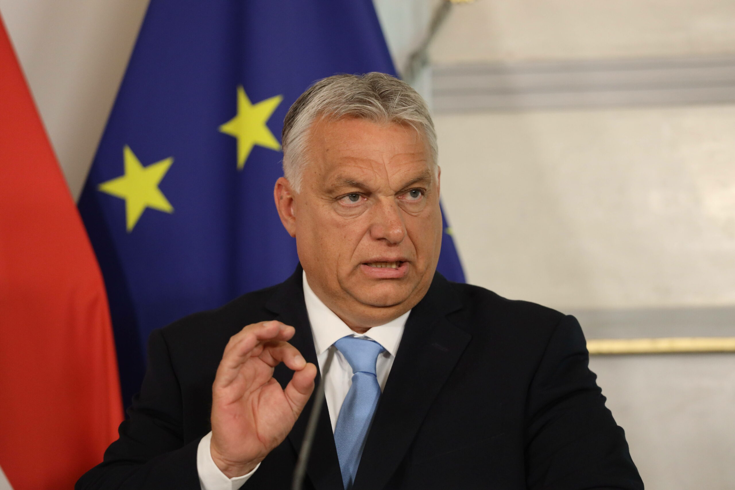 Man sieht Viktor Orban bei einer Rede. Er trägt einen schwarzen Anzug, weißes Hemd und hellblaue Krawatte, hinter ihm ist die Europäische Flagge zu sehen.