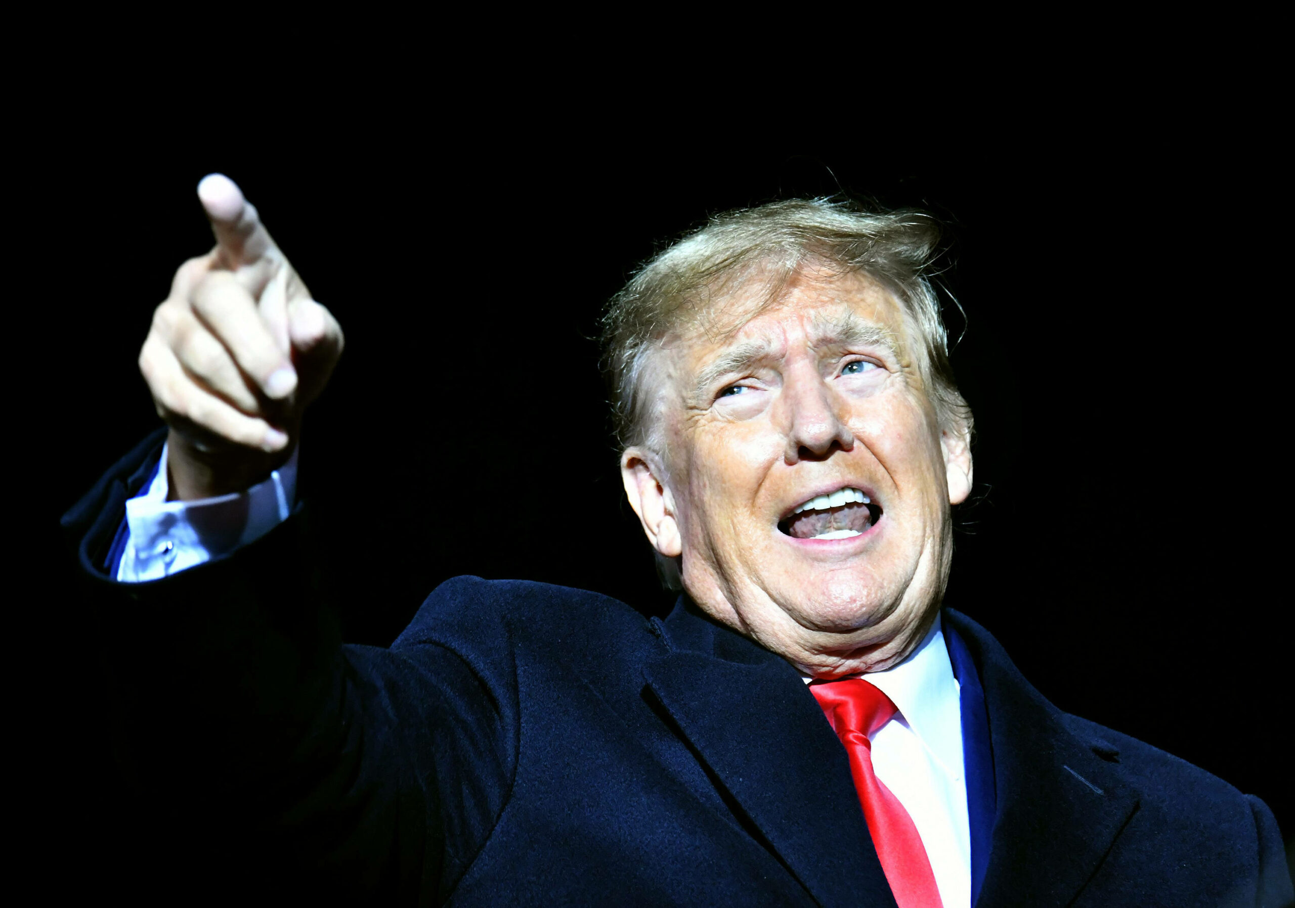 Man sieht Donald Trump wie er energisch spricht und irgendwo mit seinem rechten Zeigefinger hinzeigt. Er trägt einen schwarzen Anzug, ein weißes Hemd und eine rote Krawatte; der Hintergrund ist schwarz.