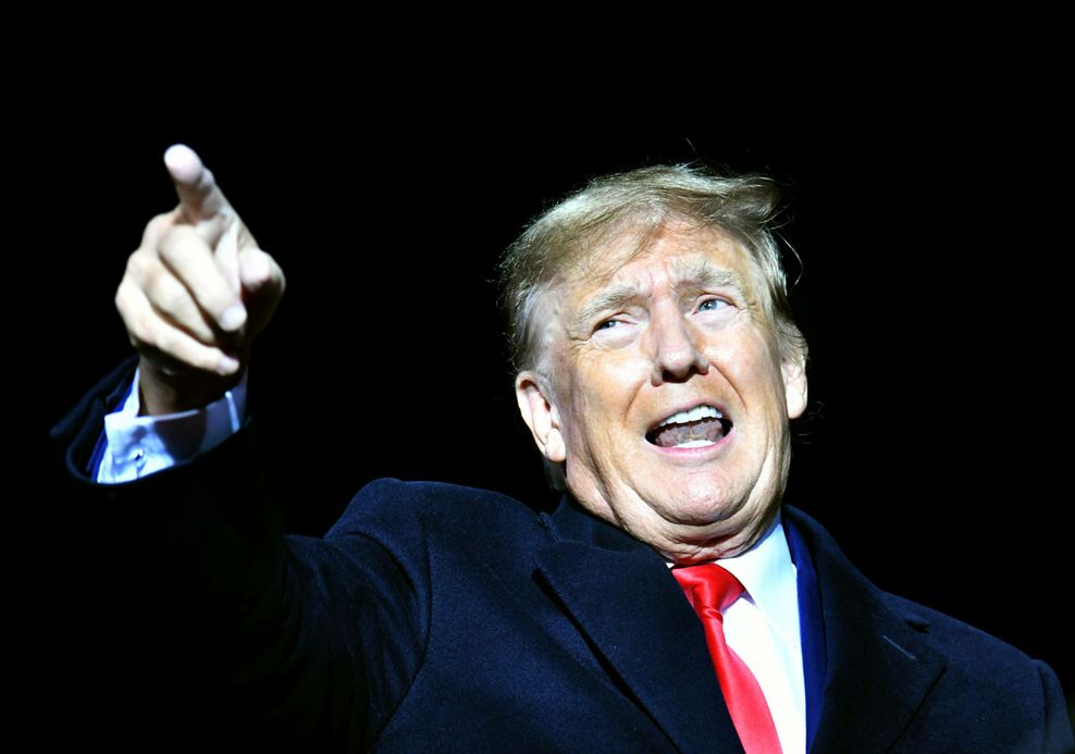 Man sieht Donald Trump wie er energisch spricht und irgendwo mit seinem rechten Zeigefinger hinzeigt. Er trägt einen schwarzen Anzug, ein weißes Hemd und eine rote Krawatte; der Hintergrund ist schwarz.